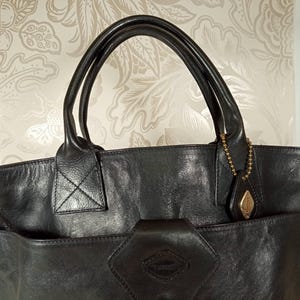 May include: Black leather tote bag with a top handle and a detachable shoulder strap. The bag has a large front pocket with a decorative metal charm.
