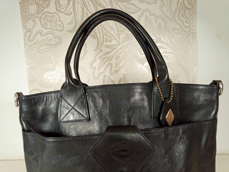 May include: Black leather tote bag with a top handle and a detachable shoulder strap. The bag has a large front pocket with a decorative metal charm.