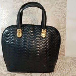May include: A black handbag featuring a quilted wave pattern across the exterior surface. The bag includes two rounded top handles attached with gold-toned metal hardware accents. The structure is rounded at the top and flat at the base.