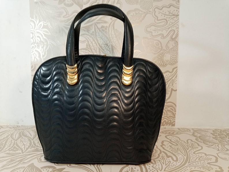 May include: A black handbag featuring a quilted wave pattern across the exterior surface. The bag includes two rounded top handles attached with gold-toned metal hardware accents. The structure is rounded at the top and flat at the base.