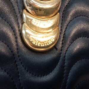 May include: A dark navy blue leather handbag featuring a quilted wave pattern. The front is adorned with a vertical row of three polished gold-tone metal crescent-shaped accents, with the word valentino engraved on the bottom metal piece.