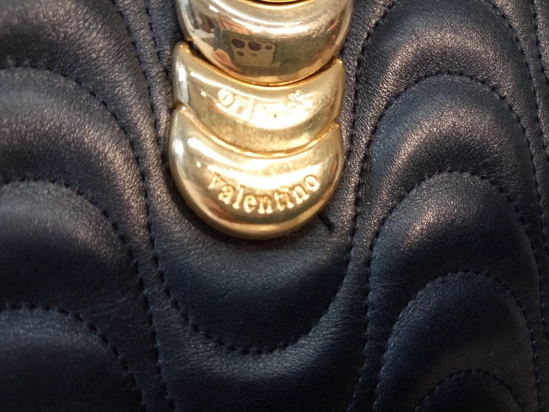 May include: A dark navy blue leather handbag featuring a quilted wave pattern. The front is adorned with a vertical row of three polished gold-tone metal crescent-shaped accents, with the word valentino engraved on the bottom metal piece.