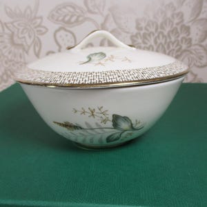 May include: A white ceramic sugar bowl with a lid, featuring a gold rim and a decorative band of gold and white squares. The bowl is adorned with green and gold floral designs. The lid has a small handle.