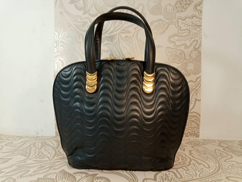May include: A black handbag featuring a quilted wave pattern across the surface. The bag has two rounded handles attached with gold-toned metal hardware. The top closure is secured with two gold-toned circular zipper pulls.