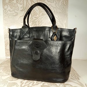 May include: A black leather tote bag with a large pocket on the front and a small metal charm attached to the handle.