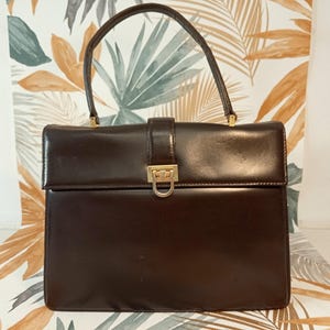 May include: A dark brown leather handbag with a rectangular shape and a top handle. The bag features a gold-colored clasp and a buckle closure. The background has a floral pattern.