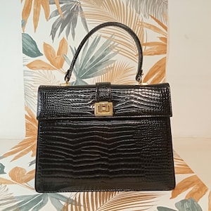 May include: Black structured handbag with a reptile-like texture. The bag features a top handle, a rectangular shape, and a gold-colored clasp. The background has a floral pattern with shades of orange, green, and white.