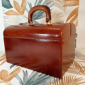 Puede incluir: Un neceser vintage de cuero marrón con la parte superior curvada y un asa resistente. El estuche tiene un ribete dorado y un cierre seguro. El asa es de color marrón oscuro. El estuche está sobre una superficie estampada.