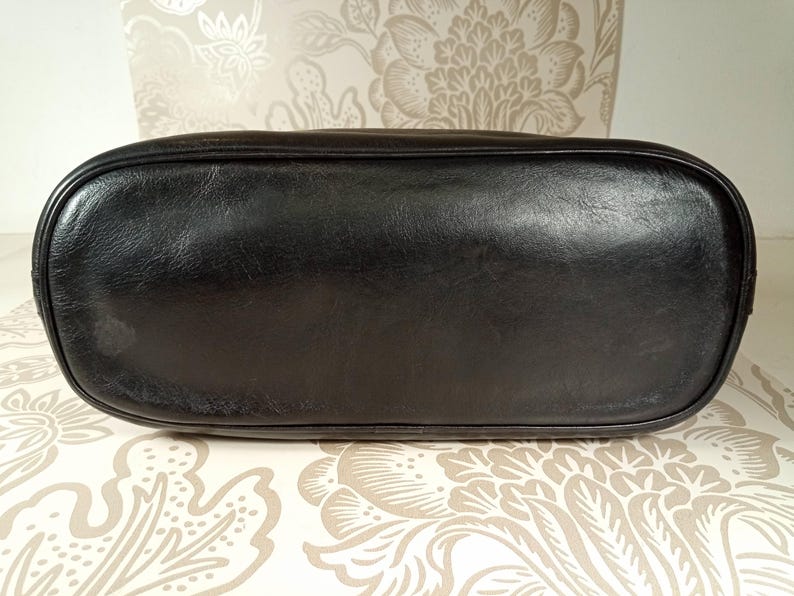 May include: Black leather tote bag with a smooth, flat bottom. The bag is empty and sitting on a patterned surface.