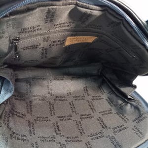 May include: The interior of a dark blue bag featuring a black fabric lining with the repeating text Valentino Orlandi. A rectangular tan leather patch is sewn inside with the text Valentino Orlandi Made in Italy. A zippered pocket is visible.