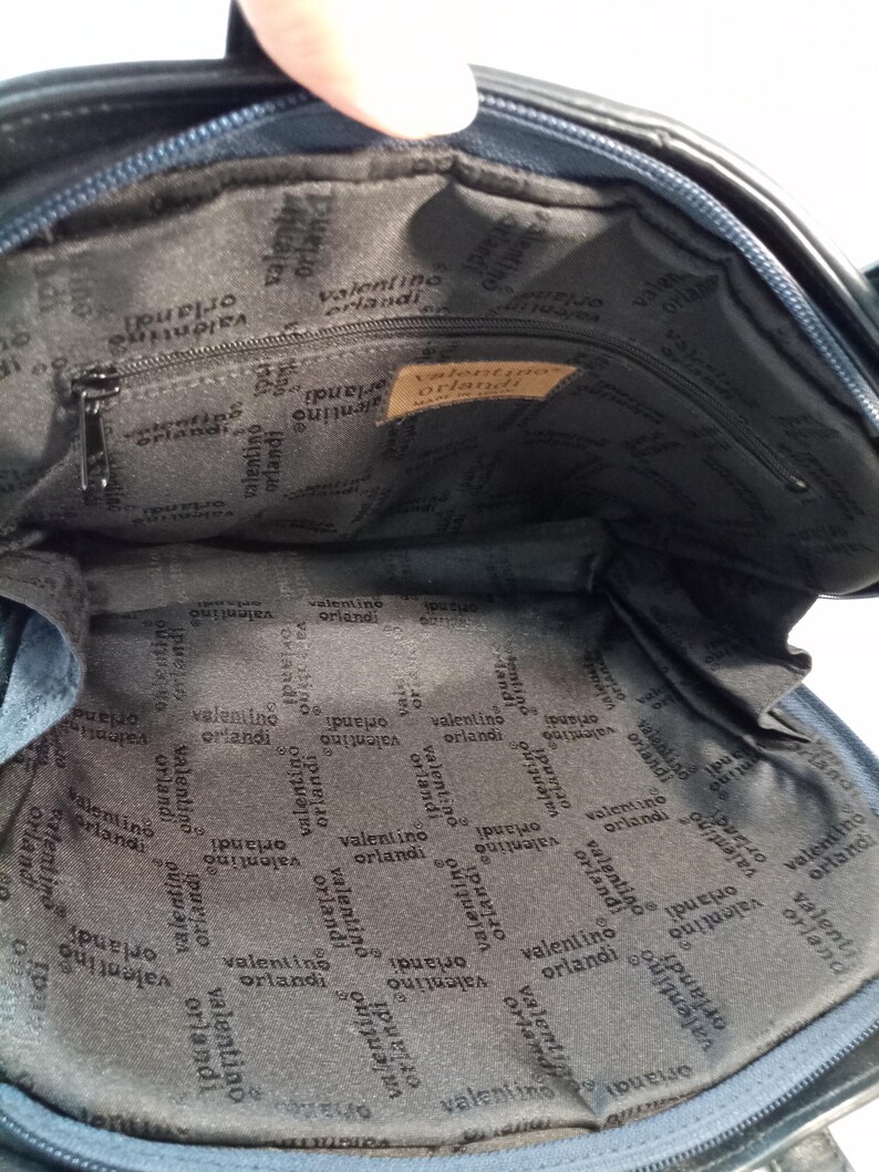 May include: The interior of a dark blue bag featuring a black fabric lining with the repeating text Valentino Orlandi. A rectangular tan leather patch is sewn inside with the text Valentino Orlandi Made in Italy. A zippered pocket is visible.