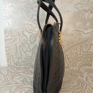 May include: A black handbag with a quilted wave pattern, featuring two tall handles and gold-toned hardware. The bag has a vertical zipper closure along the side and a structured, rounded silhouette with a smooth finish on the edges.