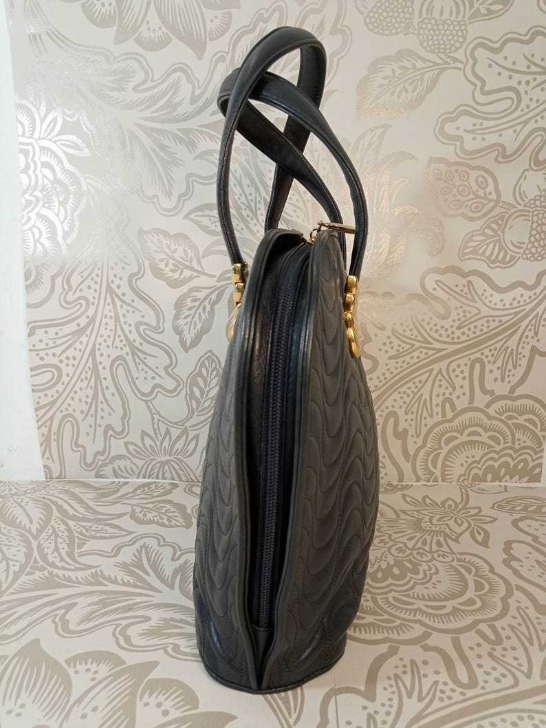 May include: A black handbag with a quilted wave pattern, featuring two tall handles and gold-toned hardware. The bag has a vertical zipper closure along the side and a structured, rounded silhouette with a smooth finish on the edges.