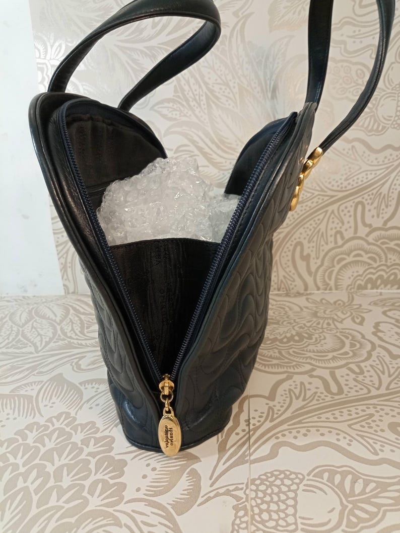 May include: A black quilted leather handbag featuring a curved silhouette, two top handles, and a gold-toned zipper pull with embossed text. The interior is partially visible, containing clear bubble wrap, and the exterior has a wavy stitched pattern.
