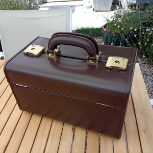 May include: A rectangular dark brown hard-shell vanity case featuring a sturdy top handle and two gold-tone metal latches. The exterior has a smooth, solid finish with rounded corners and a hinged lid design, providing a structured storage solution.