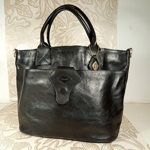 May include: A black leather tote bag with a large pocket on the front and a small metal charm attached to the handle.