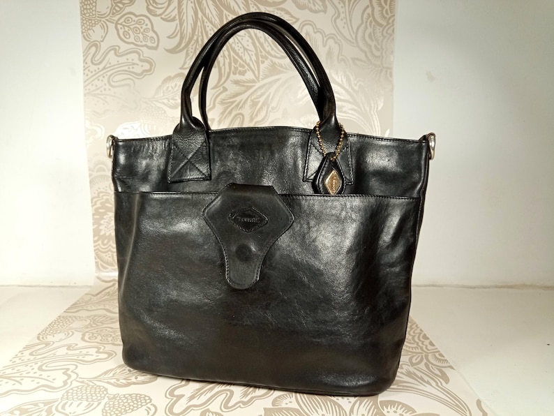 May include: A black leather tote bag with a large pocket on the front and a small metal charm attached to the handle.