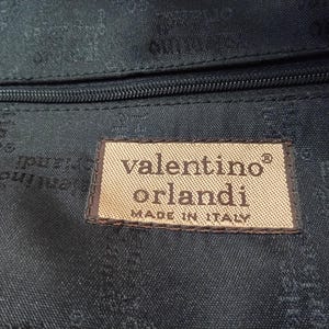 May include: A black fabric interior lining featuring a repeating Valentino Orlandi logo pattern. A rectangular beige fabric patch is stitched onto the lining, displaying the text Valentino Orlandi and MADE IN ITALY in dark brown lettering.
