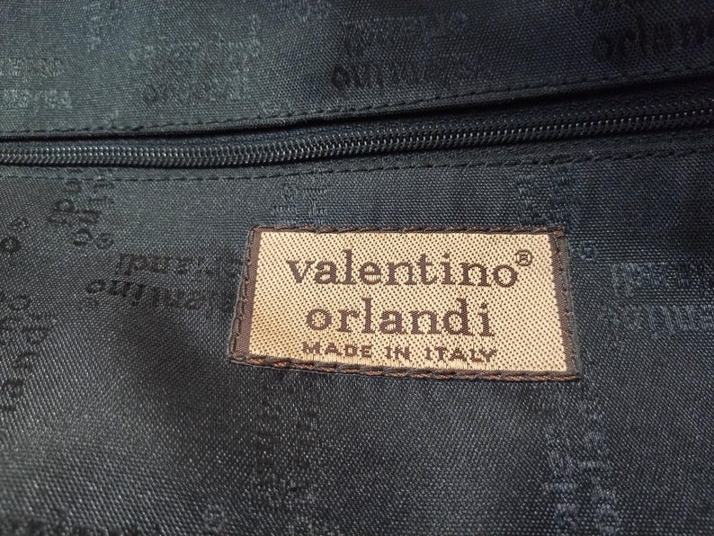May include: A black fabric interior lining featuring a repeating Valentino Orlandi logo pattern. A rectangular beige fabric patch is stitched onto the lining, displaying the text Valentino Orlandi and MADE IN ITALY in dark brown lettering.