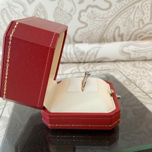 May include: A red octagonal jewelry box with gold trim, open to reveal a diamond ring on a white cushion. The box is on a glass surface, with a patterned background.