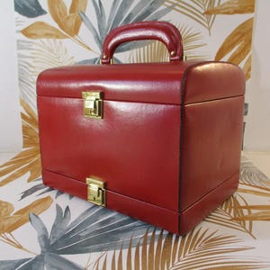 Può includere: Un portagioie in pelle rossa con manico e chiusure color oro. La scatola è rettangolare con bordi arrotondati e un manico sulla parte superiore. Lo sfondo presenta un motivo floreale.