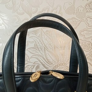 May include: A dark navy blue quilted handbag featuring two rounded leather handles and two gold-toned metal zipper pulls. The bag has a distinct stitched pattern across the surface and is positioned against a light-colored floral patterned background.