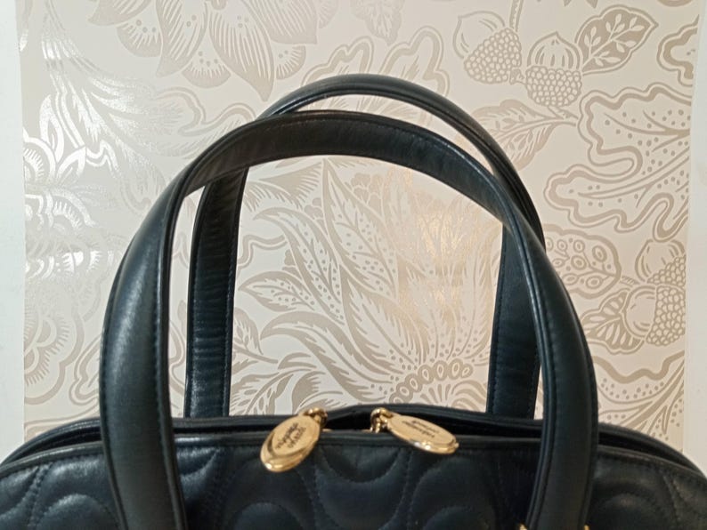 May include: A dark navy blue quilted handbag featuring two rounded leather handles and two gold-toned metal zipper pulls. The bag has a distinct stitched pattern across the surface and is positioned against a light-colored floral patterned background.