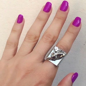 May include: A silver ring with a unique, abstract design. The ring is worn on the middle finger of a hand with purple nail polish.