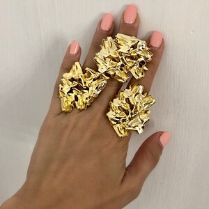 May include: Two gold-toned rings with a textured, abstract design. The rings are worn on the index and middle fingers of a hand.