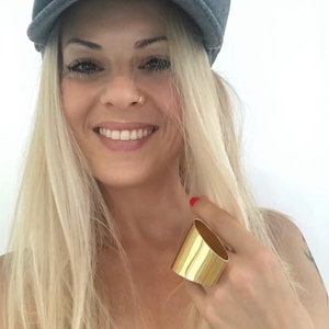May include: A woman wearing a gray newsboy cap and a gold ring on her finger. Her hair is blonde and she is smiling.