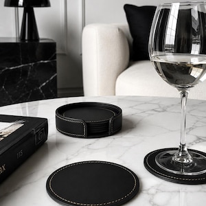 May include: A set of round black coasters with visible tan stitching along the edges. One coaster is placed under a clear glass filled with liquid, while a stack of matching coasters sits inside a circular black holder on a white marble surface.