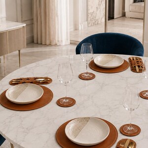 May include: A round white marble dining table set for four with tan leather placemats and matching coasters. Each place setting includes a white ceramic plate with a speckled texture, a clear glass wine goblet, and a tan leather cutlery holder.