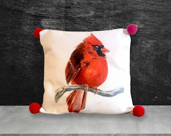 May include: A white throw pillow with a red cardinal perched on a branch. The pillow has red pom-poms on each corner.