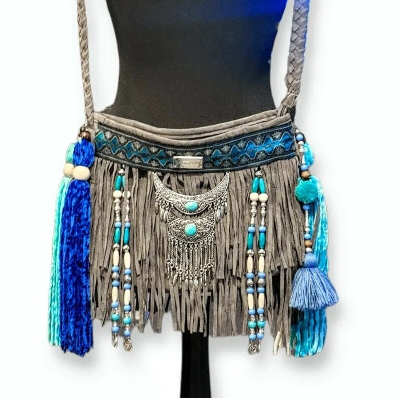 May include: A gray suede crossbody bag with blue and turquoise fringe, beads, and tassels. The bag has a silver metal pendant with turquoise stones.