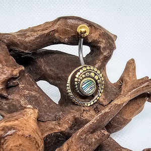 Puede incluir: Un anillo de ombligo dorado con un diseño circular de concha de abulón. El anillo está hecho de acero quirúrgico y tiene un acabado chapado en oro.