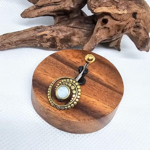 Può includere: Un piercing per ombelico dorato con un design circolare con una pietra bianca centrale e un anello esterno punteggiato. L'anello è esposto su un supporto rotondo in legno con venature naturali.