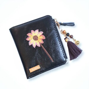 May include: Black leather wallet with a gold zip and a small embroidered flower. The wallet has a brown tassel charm with beads and a gold sun charm. The wallet has the word "Colourfabs" printed on the front.