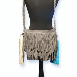 May include: Gray suede crossbody bag with fringe detailing and colorful tassels. The bag has a long adjustable strap.
