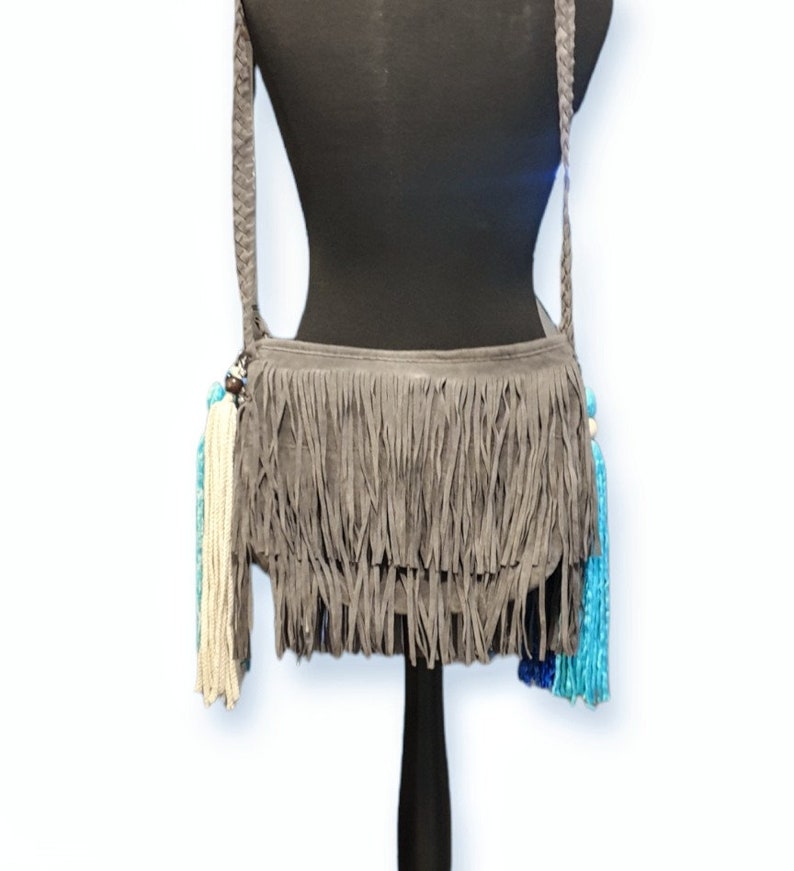 May include: Gray suede crossbody bag with fringe detailing and colorful tassels. The bag has a long adjustable strap.