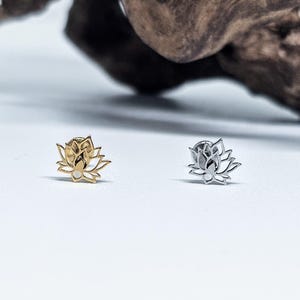 May include: Two small stud earrings, each shaped like a lotus flower. One is gold, the other silver. They sit on a white surface, with a blurred wooden background.