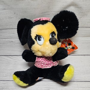May include: Vintage Minnie Mouse plush toy. The plush features black fur, a yellow face, and a pink polka dot bow and dress. The toy has a tag attached. The plush is sitting on a white wooden surface.