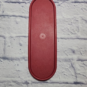 May include: A red, oval-shaped plastic lid. The lid has a slightly textured surface and a small, circular indentation in the center. The edges are rounded, and the overall shape is elongated.