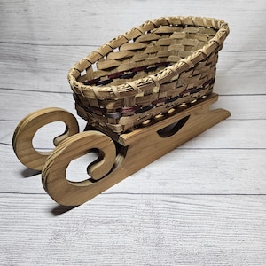 May include: A decorative wooden sled with a woven basket. The sled is made of light brown wood with curved runners. The basket is woven with natural-colored reeds and has a slightly oval shape.