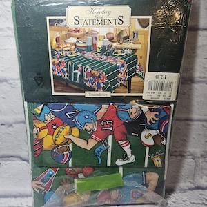 May include: A packaged oblong tablecloth with a football theme. The green tablecloth features a football field design with players in action. The packaging includes the text "Holiday Home Statements" and "Touchdown". The dimensions are 152 x 213 cm.