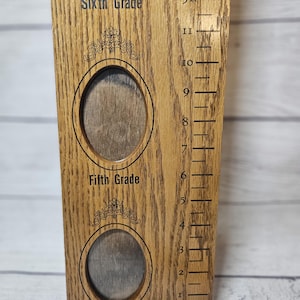 May include: Wooden growth chart with photo frames for Fourth, Fifth, and Sixth Grade. The chart has a ruler on the side with measurements in feet and inches. The text "Sixth Grade", "Fifth Grade", and "Fourth Grade" are printed on the chart.