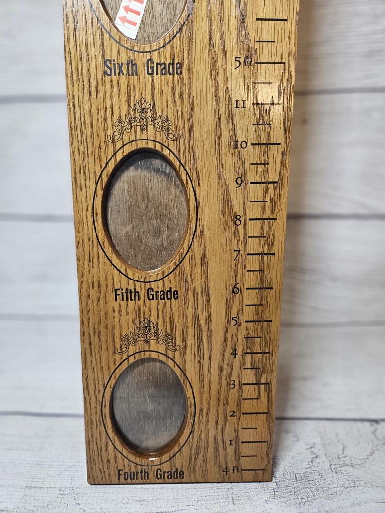 May include: Wooden growth chart with photo frames for Fourth, Fifth, and Sixth Grade. The chart has a ruler on the side with measurements in feet and inches. The text "Sixth Grade", "Fifth Grade", and "Fourth Grade" are printed on the chart.