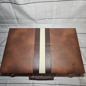 May include: A rectangular brown briefcase featuring a central vertical stripe design composed of a wide cream-colored band flanked by two dark brown strips. The case includes a brown handle and metallic latches on the bottom edge.