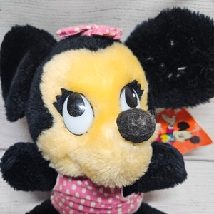 May include: A vintage Minnie Mouse plush toy with black fur, a yellow face, and a pink polka-dot bow and collar. The toy has large, expressive eyes and a black nose. A tag with a Mickey Mouse graphic is visible.