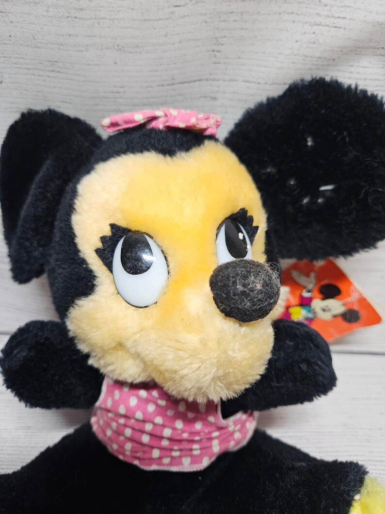 May include: A vintage Minnie Mouse plush toy with black fur, a yellow face, and a pink polka-dot bow and collar. The toy has large, expressive eyes and a black nose. A tag with a Mickey Mouse graphic is visible.