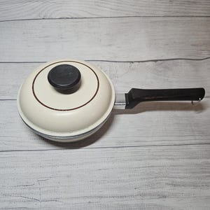 May include: A round, cream-colored saucepan with a matching lid and a long, dark brown handle. The lid features a circular dark brown knob in the center and a thin brown ring accent. The handle is attached with a silver-toned metal connector.